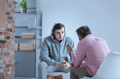 A young man in a hoodie sits and listens attentively to an older man in a pink shirt who is speaking about divorce counseling, in a room with shelves and a brick wall.